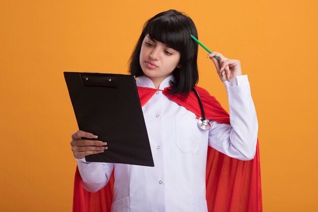 Pensando giovane ragazza di supereroi che indossa uno stetoscopio con abito medico e mantello tenendo e guardando negli appunti graffiare la testa con la matita