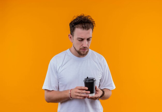 Pensando giovane ragazzo che indossa la maglietta bianca guardando la tazza di caffè sulla sua mano sulla parete arancione isolata