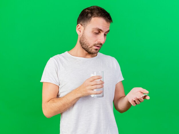 Pensando giovane uomo malato tenendo un bicchiere d'acqua e guardando le pillole in mano isolato su sfondo verde