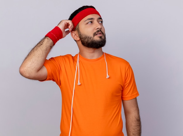 Pensando giovane uomo sportivo guardando al lato indossando la fascia e il braccialetto con le cuffie sulla spalla graffiare la testa isolata su sfondo bianco