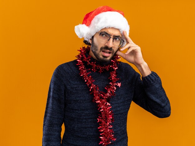 pensando guardando lato giovane bel ragazzo che indossa cappello di natale con ghirlanda sul collo mettendo la mano sulla testa isolata sul muro arancione