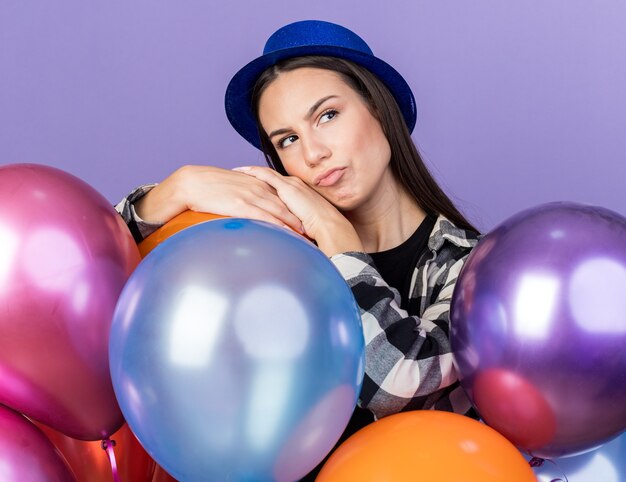 Pensando guardando lato giovane bella donna che indossa un cappello da festa in piedi dietro palloncini isolati sulla parete blu