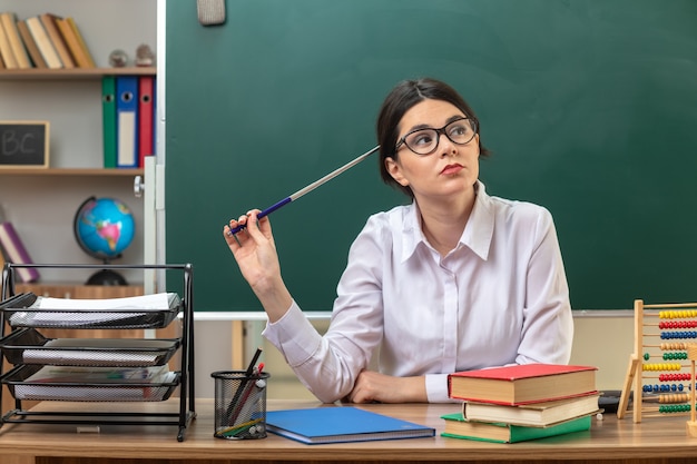 Pensando guardando lato giovane insegnante di sesso femminile con gli occhiali tenendo il puntatore stick seduto a tavola con gli strumenti della scuola in aula