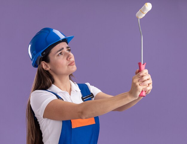 Pensando la giovane donna del costruttore in uniforme che alza e che guarda il mini rullo di vernice isolato sulla parete viola