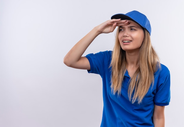 Pensando la giovane ragazza di consegna che indossa l'uniforme blu e il berretto guardando la distanza con la mano isolata su bianco