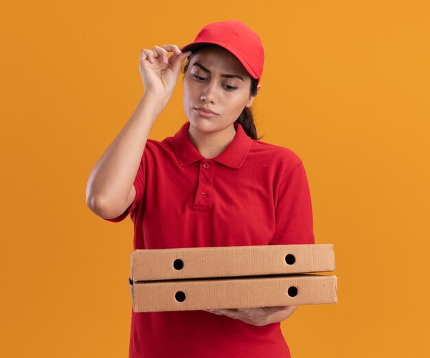 Pensando la giovane ragazza di consegna che indossa l'uniforme e la tenuta del cappuccio e guardando le scatole per pizza che tengono il cappuccio isolato sulla parete arancione