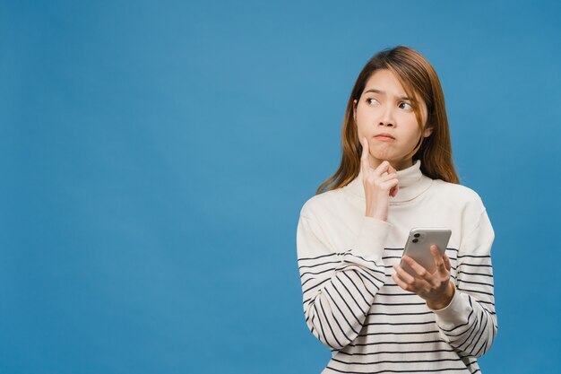 Pensare di sognare una giovane donna asiatica che usa il telefono con espressione positiva, vestita con abiti casual, sentendo felicità e stando isolata sul muro blu