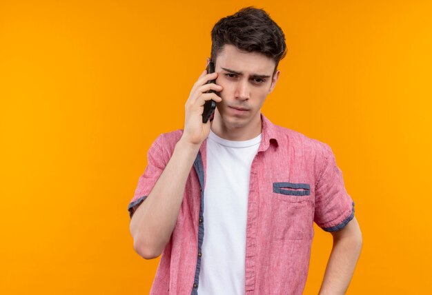 Pensare il giovane ragazzo caucasico che indossa la camicia rosa parla al telefono sulla parete arancione isolata