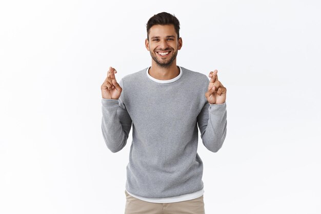 Pensiero positivo, preghiera e concetto di credenza Fiducioso sorridente ottimista bel ragazzo in maglione grigio incrociare le dita per buona fortuna anticipando buone notizie assaporare in attesa di buon affare sfondo bianco