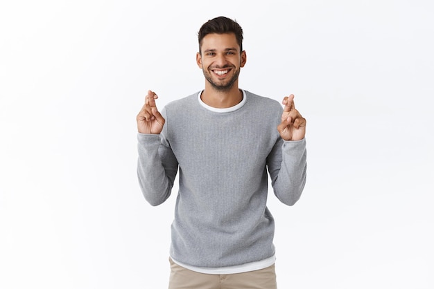 Pensiero positivo, preghiera e concetto di credenza Fiducioso sorridente ottimista bel ragazzo in maglione grigio incrociare le dita per buona fortuna anticipando buone notizie assaporare in attesa di buon affare sfondo bianco