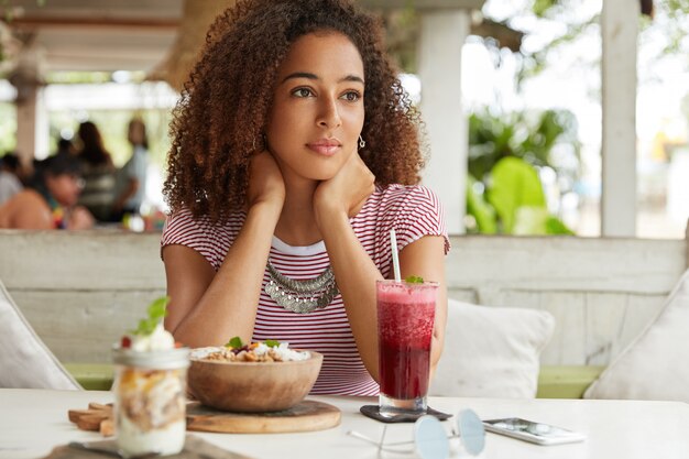 Pensierosa adorabile giovane donna afroamericana ricrea al bar con cocktail esotici e insalata, pensa ai programmi nei fine settimana, essendo immersa nei pensieri. Persone, etnia e concetto di relax