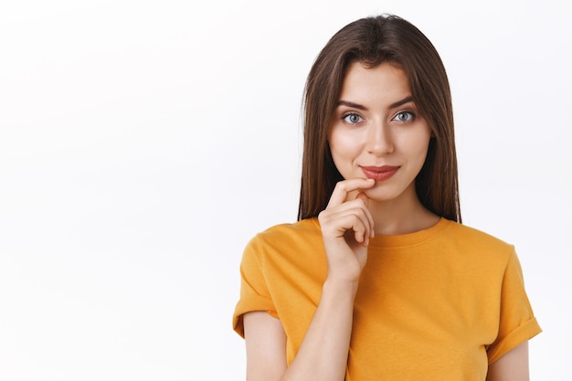 Pensieroso bella audace giovane donna moderna in maglietta gialla sorridente sfacciata e creativa, toccando il labbro ha un'idea interessante, incuriosita dal promo, in piedi sfondo bianco pensando