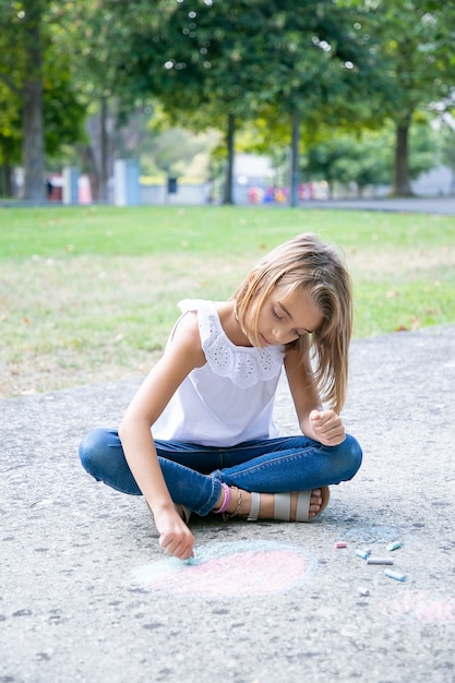 Pensieroso bella ragazza seduta e disegno con pezzi colorati di gessi. Vista frontale. Concetto di infanzia e creatività