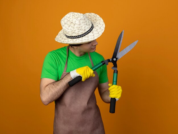 pensieroso giovane giardiniere maschio in uniforme che indossa il cappello da giardinaggio con guanti tenendo e guardando clippers isolato su arancione