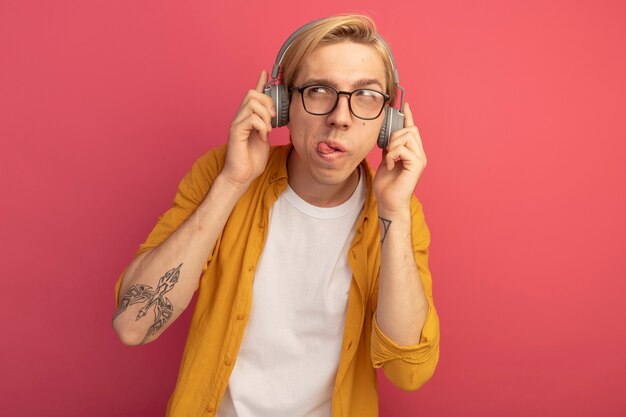 pensieroso guardando al lato giovane ragazzo biondo che indossa t-shirt gialla e occhiali con le cuffie che mostrano la lingua isolata sul rosa con spazio di copia