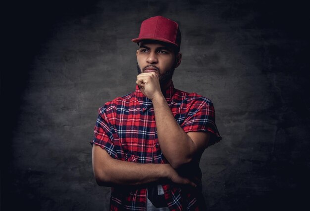 Pensieroso un ragazzo hipster afroamericano vestito con una camicia di pile rossa e un berretto in studio. Isolato su uno sfondo scuro con texture.