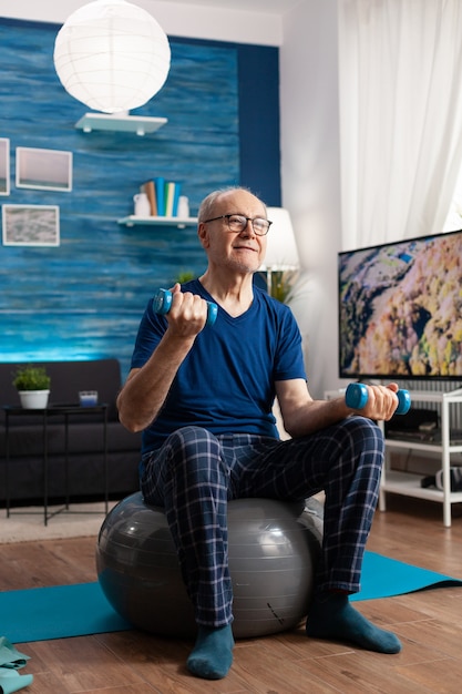 Pensionamento uomo anziano seduto sulla palla svizzera che esercita i muscoli delle braccia facendo esercizi di fitness utilizzando manubri di allenamento. Pensionato focalizzato che allena la resistenza della forza del corpo in soggiorno