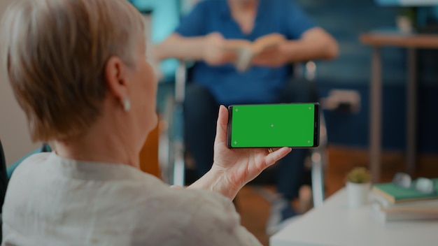 Pensionato femminile che esamina lo schermo verde orizzontale sul telefono cellulare. Vecchia donna con le stampelle che tiene lo smartphone con la chiave cromatica vuota e lo sfondo dello spazio di copia isolato. Modello di simulazione