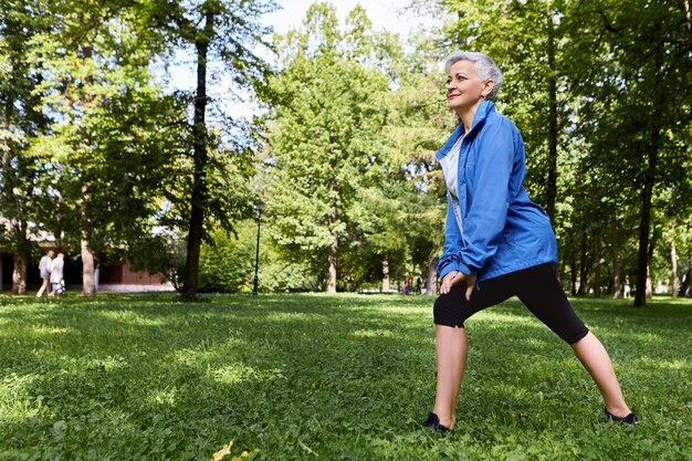 Pensionato femminile energico in abbigliamento sportivo alla moda scegliendo un allenamento sano stile di vita attivo sull'erba verde nella foresta o nel parco, facendo affondi, avendo uno sguardo gioioso felice. Persone anziane, fitness ed estate