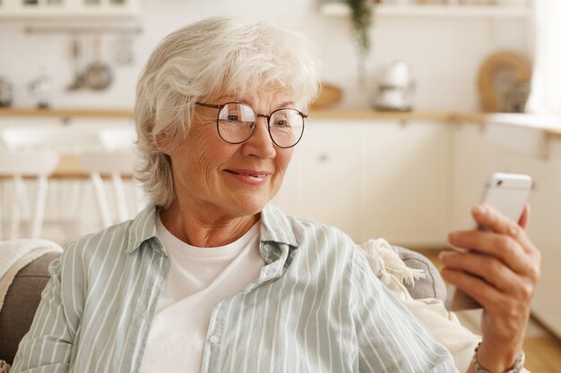 Pensionato femminile gioioso felice in occhiali rotondi navigando in internet sul cellulare, guardando lo schermo del cellulare con un ampio sorriso, prenotando biglietti aerei, pianificando un viaggio o scorrendo foto tramite social network