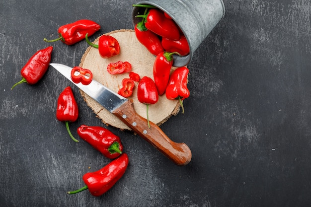 Peperoni sparsi con pezzo di legno, coltello in un mini secchio sul muro grigio sgangherata, piatto laici.