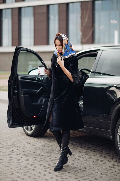 Per tutta la lunghezza di una signora alla moda in cappotto nero e stivali di pelle che indossa un fazzoletto colorato intorno al collo. Sta portando una tazza di caffè da asporto contro l'auto aperta in strada.
