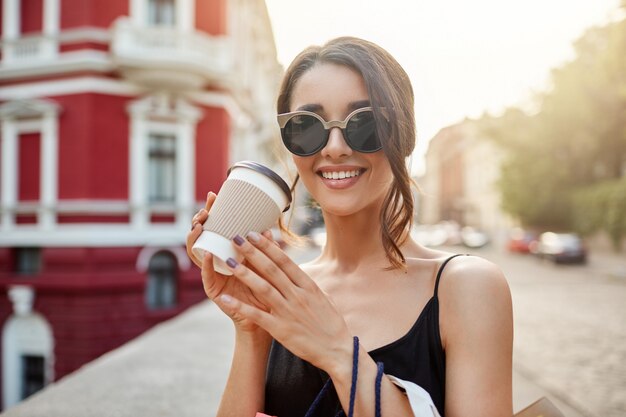 perdere il ritratto della giovane e bella ragazza caucasica con i capelli scuri in occhiali da sole e abito nero sorridente con i denti, bere caffè, rilassarsi dopo un lungo shopping nel centro commerciale.