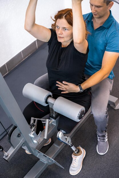 Personal trainer che assiste la donna con disabilità nel suo allenamento. Centro di riabilitazione sportiva con fisioterapisti e pazienti che lavorano insieme per la guarigione.