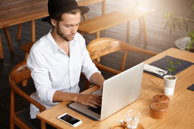 Persone, affari e concetto di tecnologia moderna. Uomo alla moda in black hat tastiera sul laptop generico, utilizzando la connessione internet ad alta velocità, seduto al tavolo del caffè durante la pausa caffè