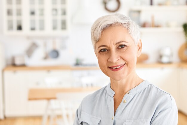 Persone anziane, maturità e concetto di pensionamento. Affascinante pensionato donna europea allegra vestita in abiti casual godendo calma mattina al chiuso, essere di buon umore, aspettando il figlio per la colazione