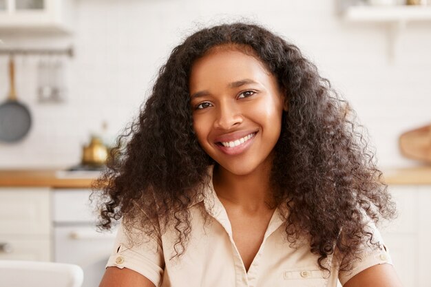 Persone, bellezza, etnia, femminilità ed emozioni umane positive. Carina bella giovane femmina africana con affascinante sorriso raggiante, rallegrandosi di buone notizie, facendo colazione in cucina accogliente