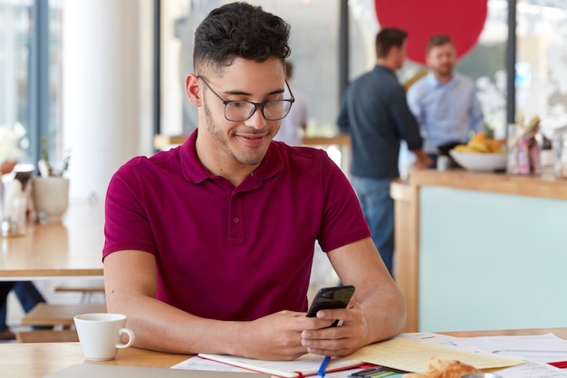 Persone, carriera, concetto di tecnologia. Il bello hipster maschio con gli occhiali tiene il cellulare, gode della comunicazione online, connesso al 4G, impegnato con le scartoffie alla caffetteria, beve caffè aromatico