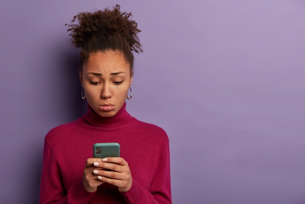 Persone, concetto di emozioni infelici. La donna triste e delusa ha ricevuto un sms negativo, tiene lo smartphone e si concentra sullo schermo con insoddisfazione, indossa poloneck bordeaux, posa al coperto, si sente solo