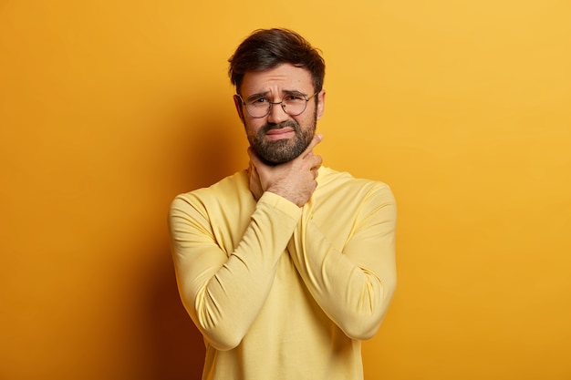 Persone, concetto di problemi di salute. L'uomo infelice e frustrato soffre di mal di gola, tocca il collo con le mani, sembra insoddisfatto, indossa occhiali rotondi e maglione giallo, ha un attacco d'asma