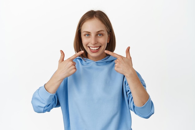 Persone dal dentista. Donna caucasica felice sorridente che mostra il suo sorriso bianco perfetto, indicando i denti e guardando gioiosa, in piedi in felpa con cappuccio blu su sfondo bianco.