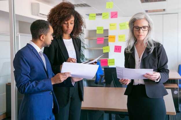 Persone di affari focalizzate che leggono documenti con statistiche. Datori di lavoro di ufficio concentrati di successo in giacca e cravatta che si incontrano nella stanza dell'ufficio e studiano i rapporti. Concetto di lavoro di squadra, affari e gestione