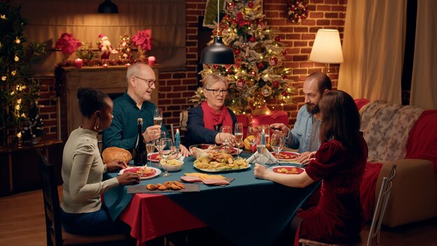 Persone diverse che ridono si sono radunate intorno al tavolo della cena di Natale facendo tintinnare bicchieri di vino. Felici sorridenti cordiali membri della famiglia che celebrano le tradizionali vacanze invernali a casa.