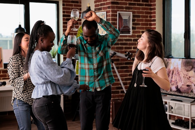 Persone diverse e felici che ascoltano musica da discoteca funky mentre bevono vino e si divertono insieme. Gruppo multietnico di amici intimi che ballano alla festa mentre celebrano un evento importante.