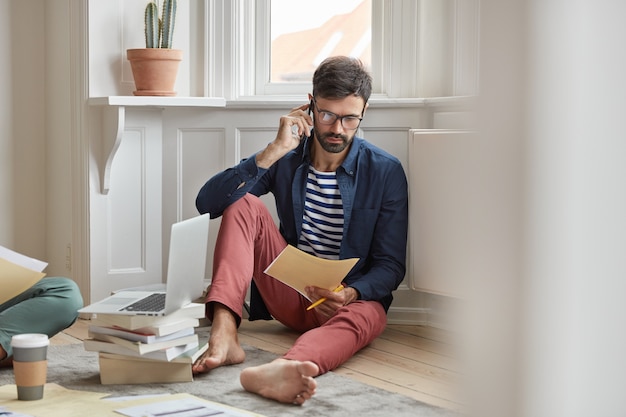 Persone e concetto di affari. Un dipendente maschio con la barba lunga pensa a una soluzione migliore, parla tramite smartphone, legge la documentazione