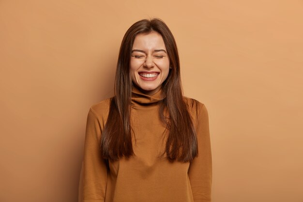 Persone e concetto di gioia. Felicissima donna adulta dai capelli scuri ride felicemente con gli occhi chiusi, parla casualmente con un amico, non può trattenere le risate, indossa un dolcevita casual, isolato sul muro marrone