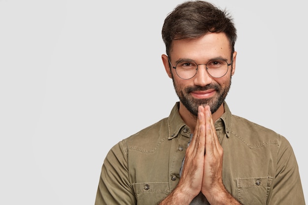 Persone e concetto di meditazione. Fedele soddisfatto giovane europeo con barba e baffi, tiene insieme i palmi delle mani, crede in qualcosa di buono, vestito con una camicia alla moda, posa da solo al coperto