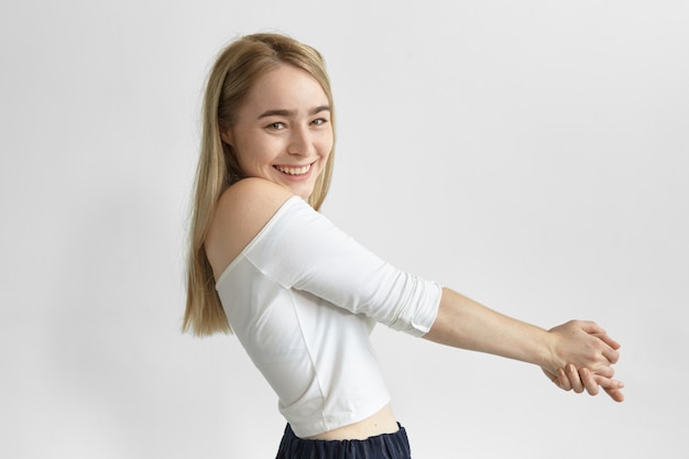 Persone e concetto di stile di vita. Colpo laterale di splendida adorabile giovane donna europea con i capelli biondi dritti in posa con le braccia tese in avanti, sorridente ampiamente, sentendosi felice