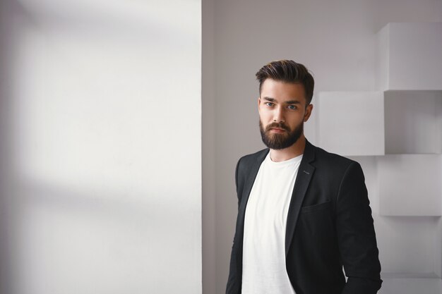 Persone e concetto di stile di vita. Elegante giovane brunetta con la barba lunga uomo d'affari vestito con elegante giacca sopra t-shirt bianca in posa isolata dalla finestra, guardando la telecamera con grave espressione facciale