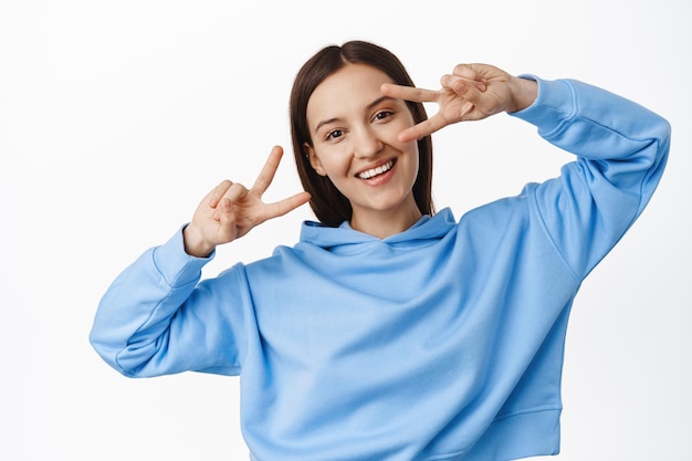 Persone e stile di vita. Giovane donna felice in felpa con cappuccio blu, che mostra segni di pace v, gesto da discoteca vicino agli occhi, ballando e sorridendo gioiosa, in piedi su sfondo bianco