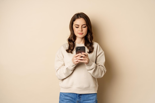 Persone e tecnologia cellulare. Bella donna alla moda che utilizza il telefono cellulare, l'app per smartphone, sorride e guarda lo schermo, in piedi su sfondo beige