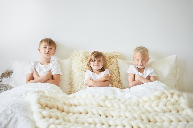 Persone, famiglia e concetto di infanzia. Tre bambini seduti uno accanto all'altro sul grande letto bianco con le braccia conserte, guardando i cartoni animati la mattina del fine settimana. Due fratelli e sorelle che giocano in casa