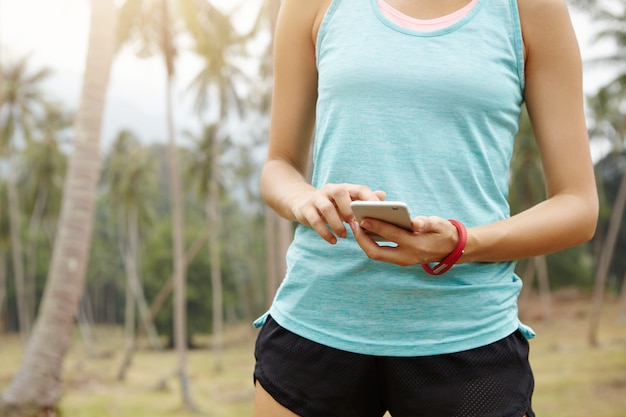 Persone, fitness e concetto di tecnologia. Sezione mediana del corridore femminile in abbigliamento sportivo utilizzando il telefono cellulare, controllando le impostazioni sull'app per monitorare i suoi progressi.