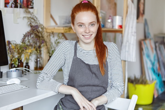 Persone, hobby, lavoro creativo e concetto di occupazione. Ritratto di giovane fiorista femminile dai capelli rossi affascinante sorridente adorabile che si rilassa nella sua officina con i fiori secchi