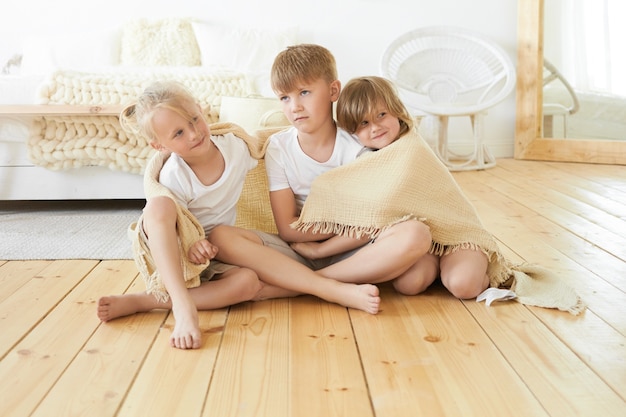 Persone, infanzia, famiglia, amore e concetto di solidarietà. Immagine dolce e accogliente di tre fratelli germani piccoli svegli che si siedono sul pavimento di legno insieme avvolti nella coperta, che si abbracciano