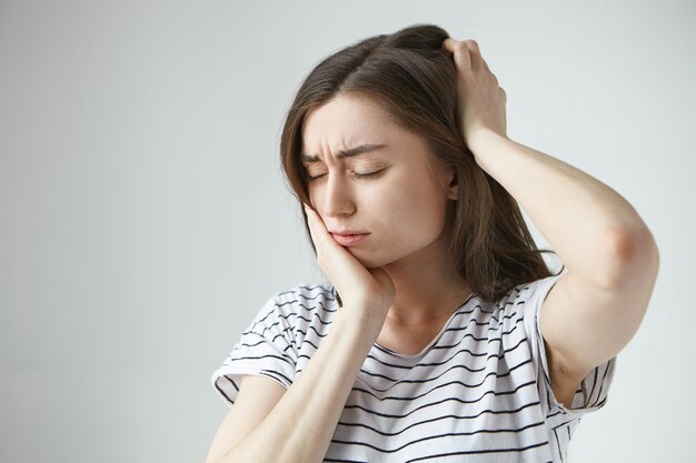 Persone, malattia e concetto di cattiva salute. Foto di infelice frustrato giovane donna bruna chiudendo gli occhi, toccando la guancia e la parte posteriore della testa mentre soffre di un terribile mal di denti